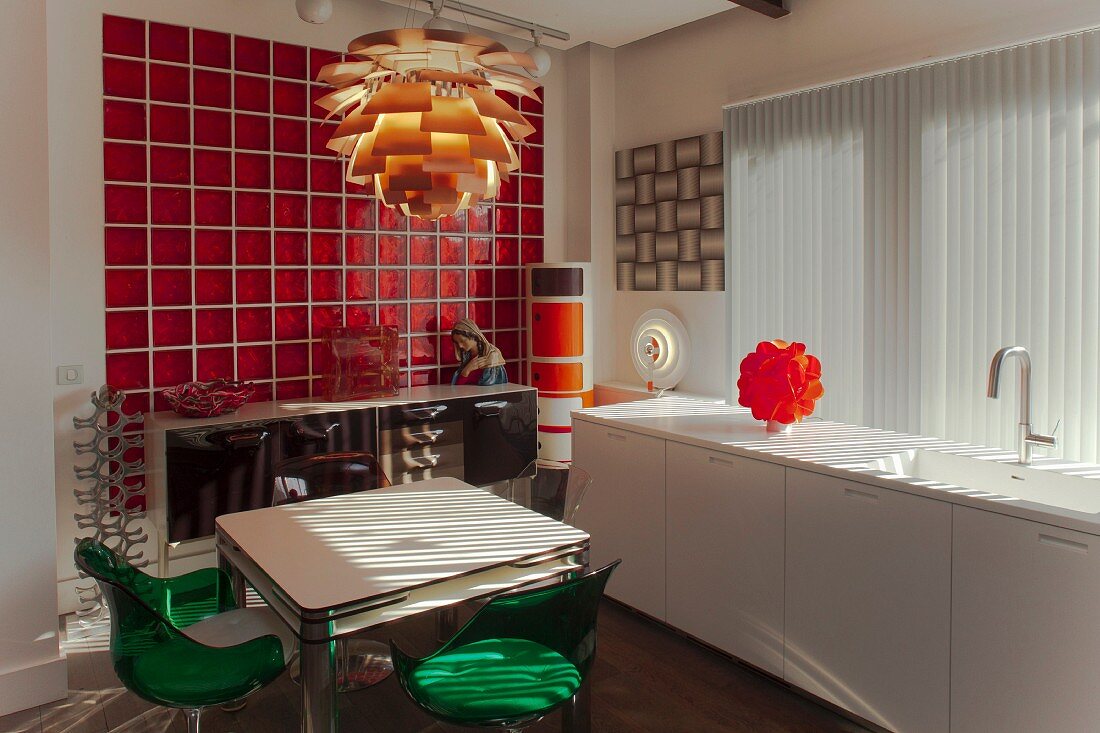 weiße Küchenzeile vor Lamellenvorhang, Essplatz unter Klassiker-Pendelleucht und Retro-Sideboard vor rote Glasfliesenwand