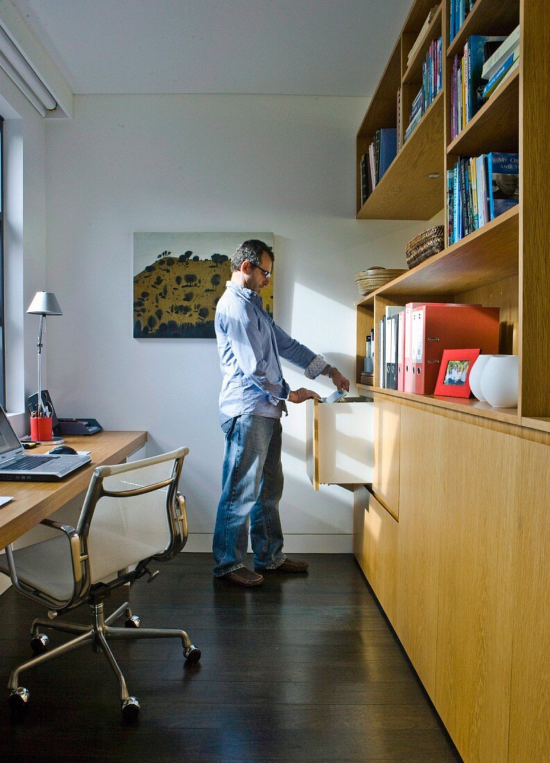 Man in narrow study with classic office chair at desk and fitted wooden cupboards