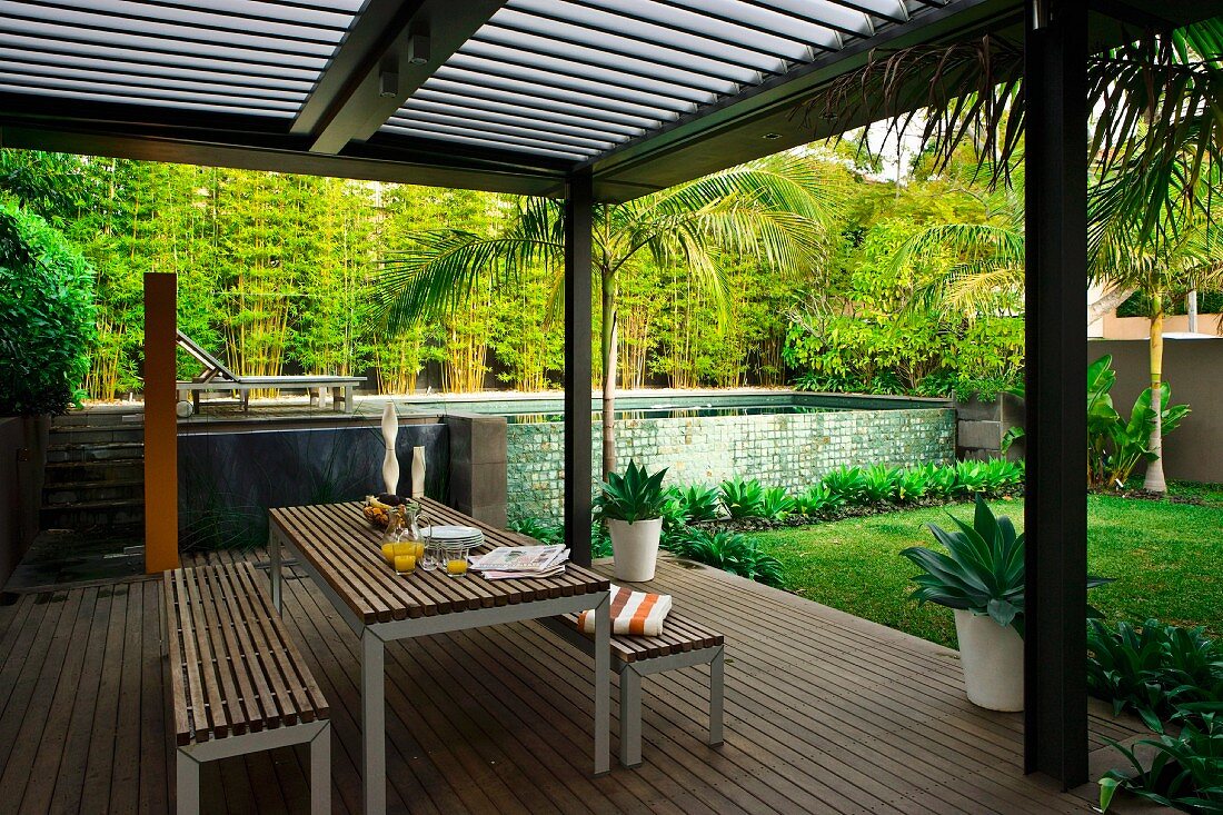 Table and bench set on modern wooden terrace below slatted pergola and pool in garden in background
