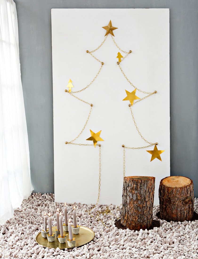 Hand-crafted stylised Christmas tree made from gold chain, stars and white plywood board behind tree stump stools and candles