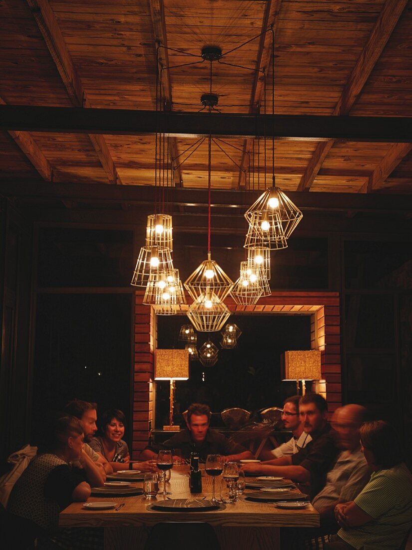 Gäste um Tisch unter beleuchteten Draht-Pendelleuchten in gemütlicher Stimmung in Safari-Lodge