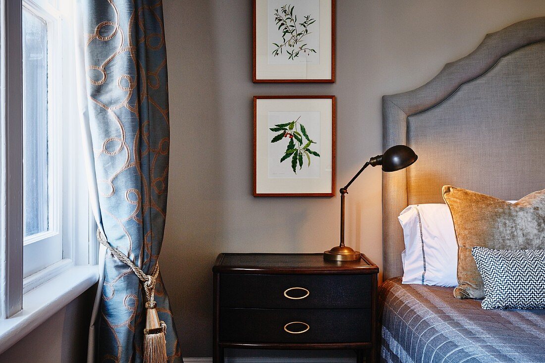 Retro Tischleuchte auf Nachtkästchen neben Bett mit gepolstertem Kopfteil, an Wand gerahmte Bilder mit Pflanzenmotiven