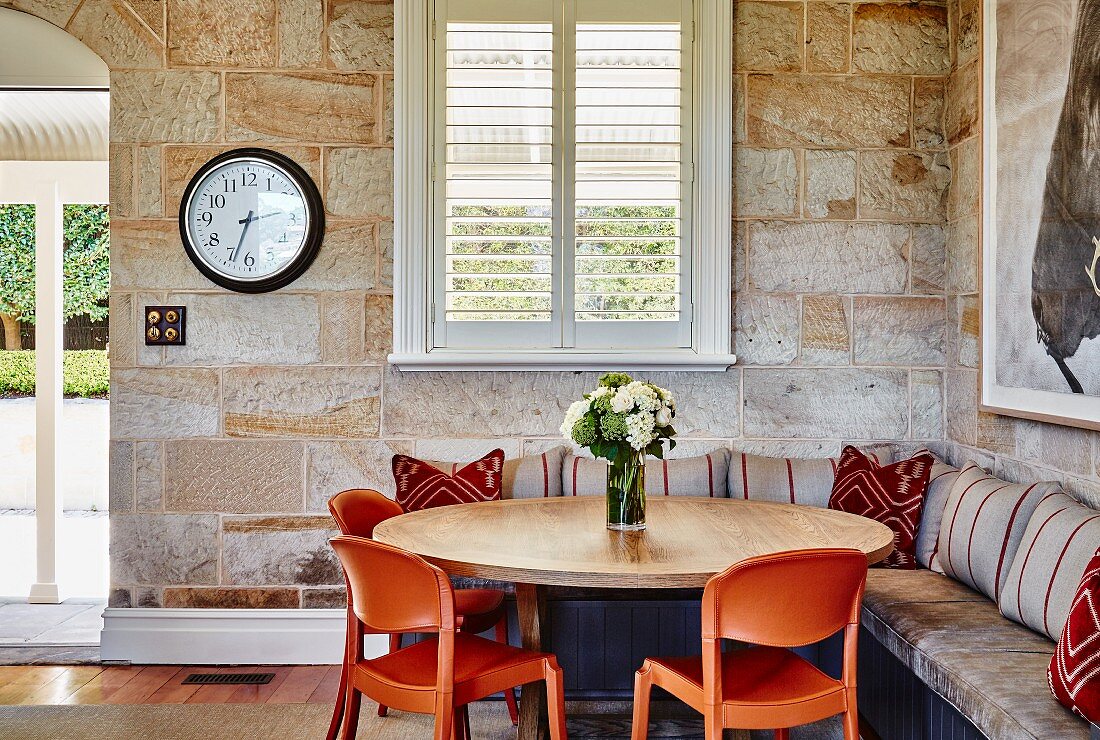Um rundem Holztisch orangefarbenen Kunststoff Stühle und eingebaute Eckbank an Natursteinwand in ländlicher Wohnzimmerecke