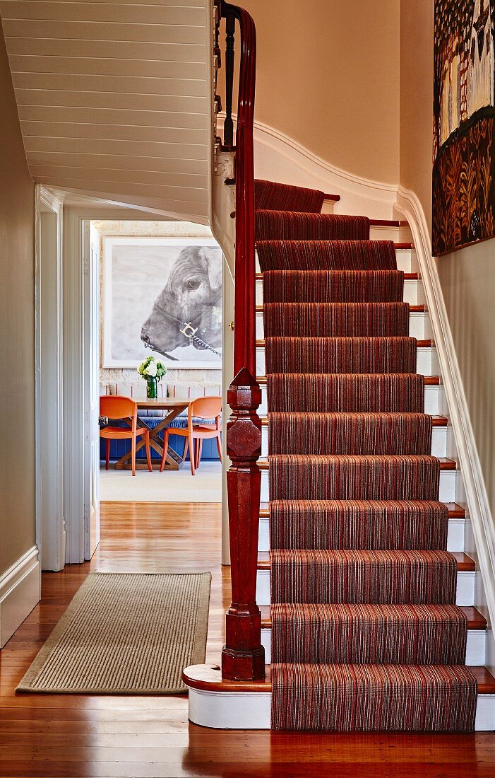 Striped runner on white-painted wooden staircase in rustic hallway; open door with view of dining area in background