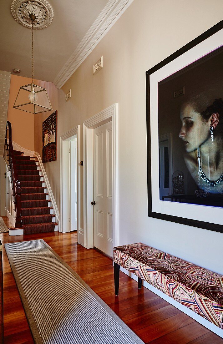 In traditionellem, elegantem Flur gepolsterte Sitzbank vor Wand mit Portrait Foto einer Frau, im Hintergrund Treppenaufgang