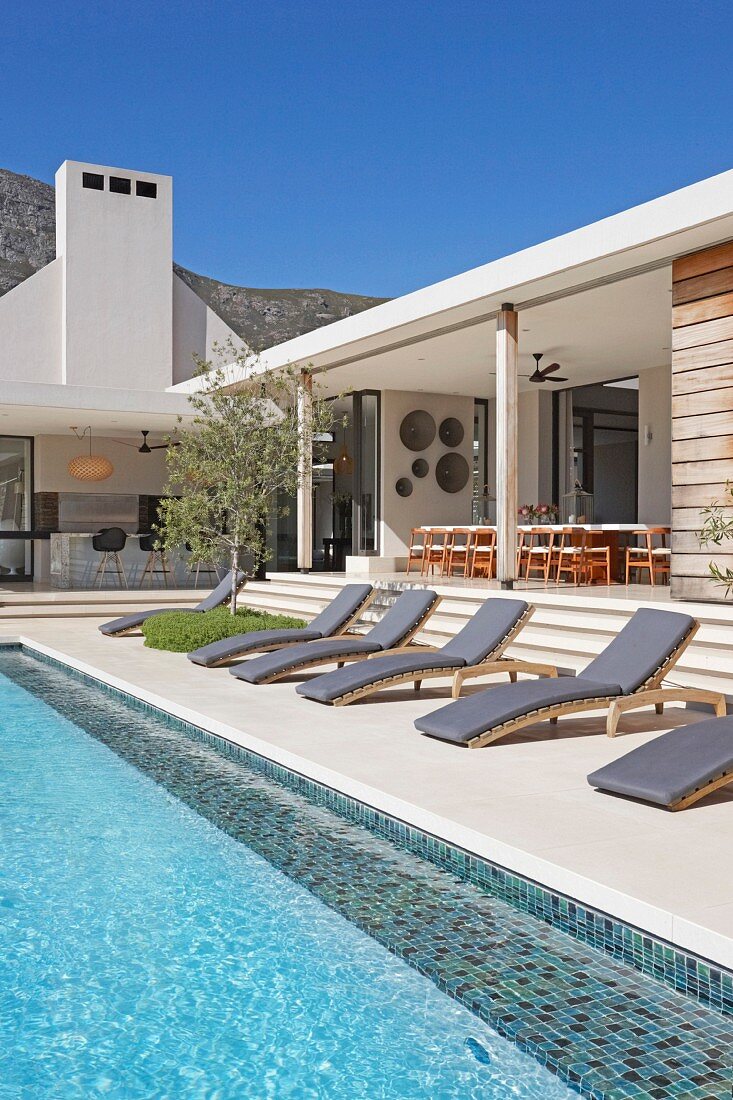 Sun loungers with grey mattresses next to pool adjoining white bungalow with spacious loggia