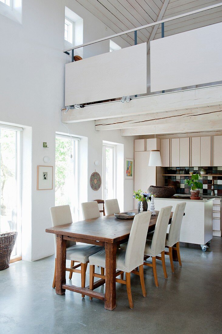 Essplatz mit Holztisch und hell gepolsterten Stühlen in offener Küche, darüber weiße Galeriebrüstung