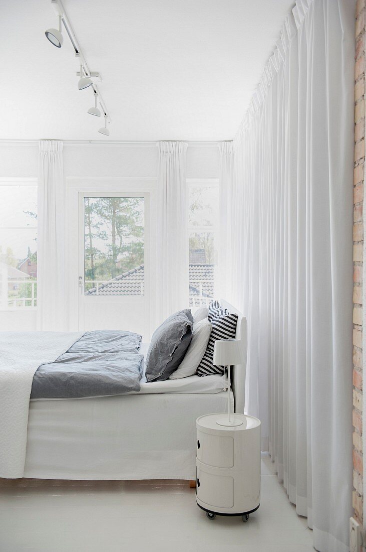 Bett mit grauer Bettwäsche und rundem Rollcontainer als Nachtkästchen vor bodenlangem Vorhang in weißem Schlafzimmer