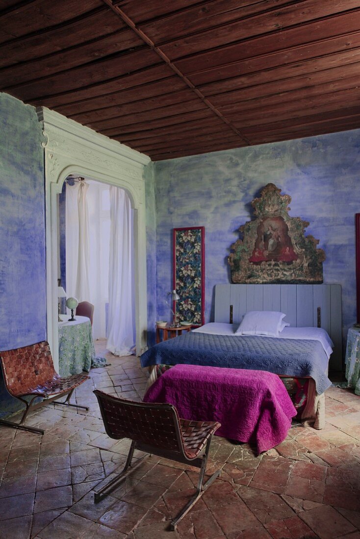 Rustic Mediterranean bedroom with religious painting on blue-painted wall, double bed and retro leather chairs