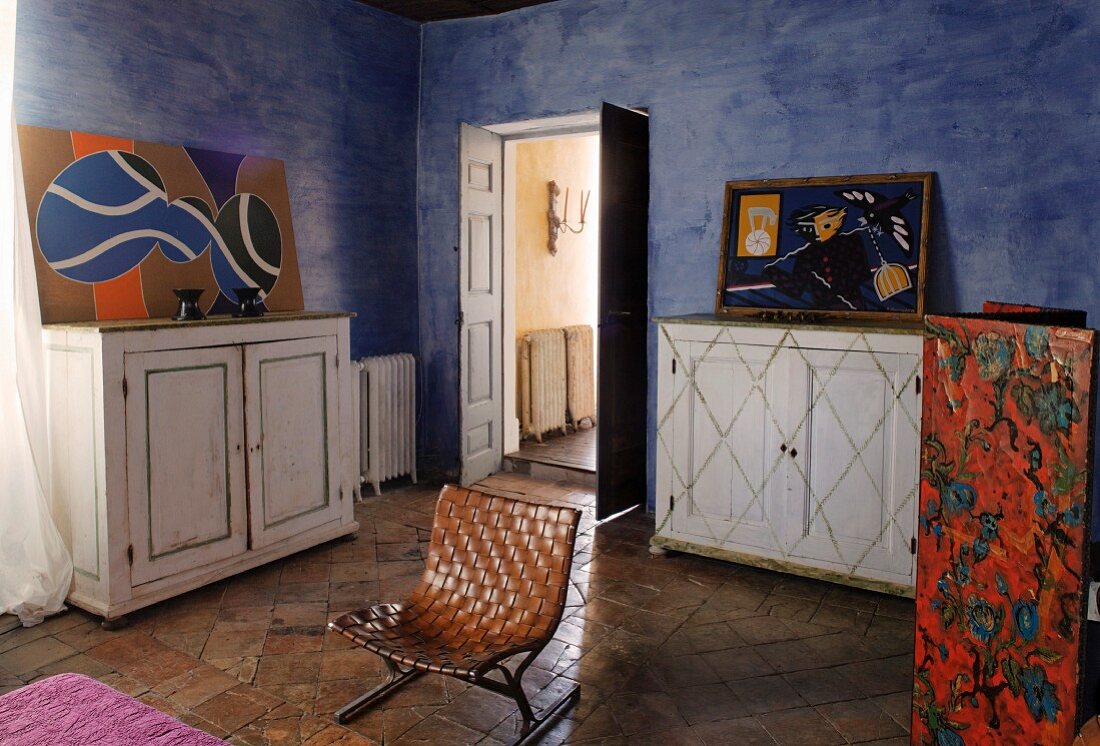 Retro-Stuhl mit Ledergeflecht zwischen weißen Vintage Sideboards in mediterranem Schlafraum mit blau getönter Wandgestaltung und modernen Bildern