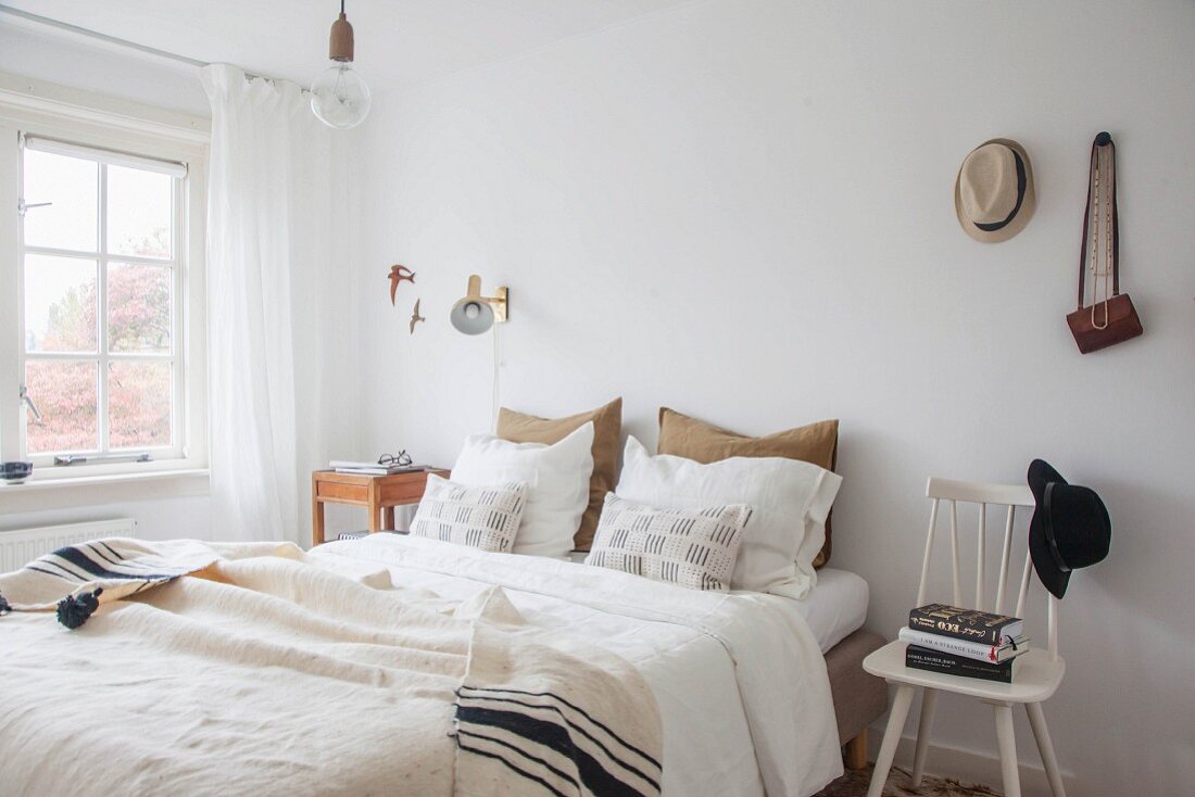 Doppelbett mit Kissen und Tagesdecke, weißer Stuhl als Nachttisch mit Bücherstapel und aufgehängtem Hut in schlichtem Schlafzimmer