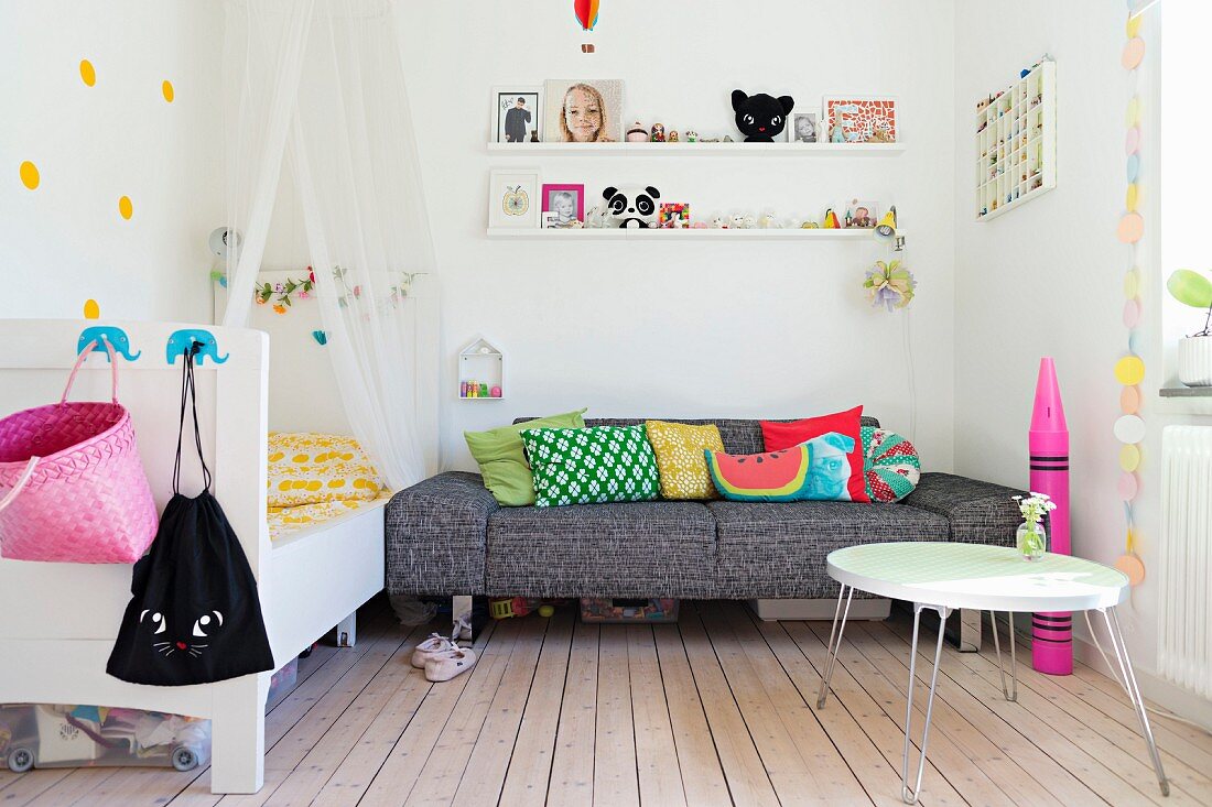 Mädchenzimmer mit Couchtisch und graumeliertem Sofa, weißen Wandboards mit Spielsachen und Bett mit Baldachin