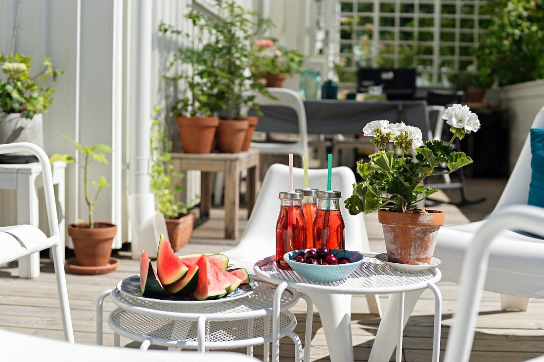 Wassermelone, Erfrischungsgetränke und Geranientopf auf mehrteiligem weißem Beistelltisch-Set auf der Terrasse