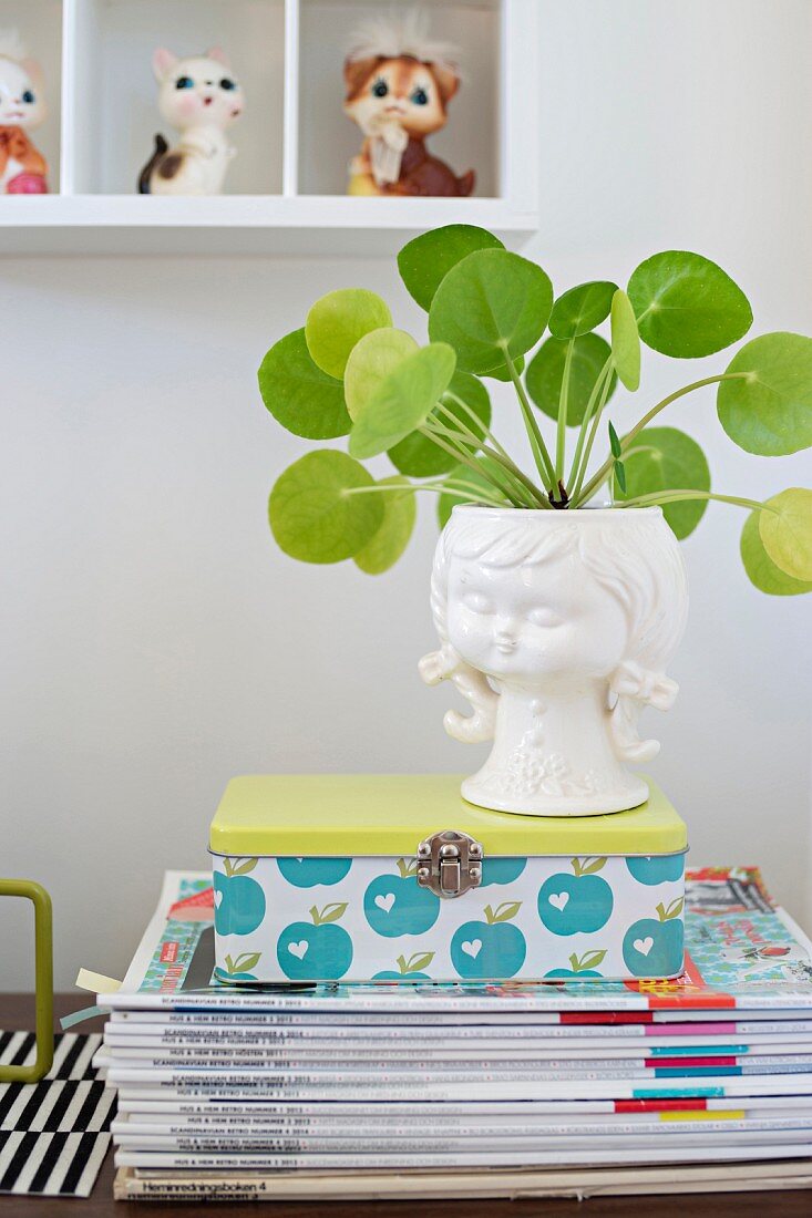 Grünpflanze in weißem Porzellanübertopf mit Mädchenmotiv auf Retro-Metalldose und Zeitschriftenstapel