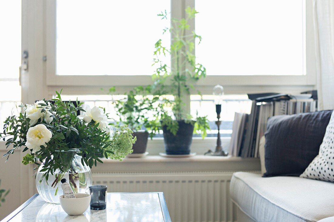 Glass vase of white flowers on table in front of house plants on windowsill