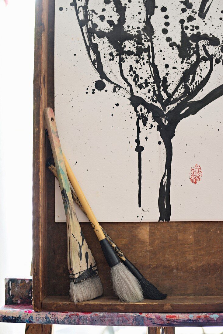 Various paintbrushes in front of black and white painting in wooden frame