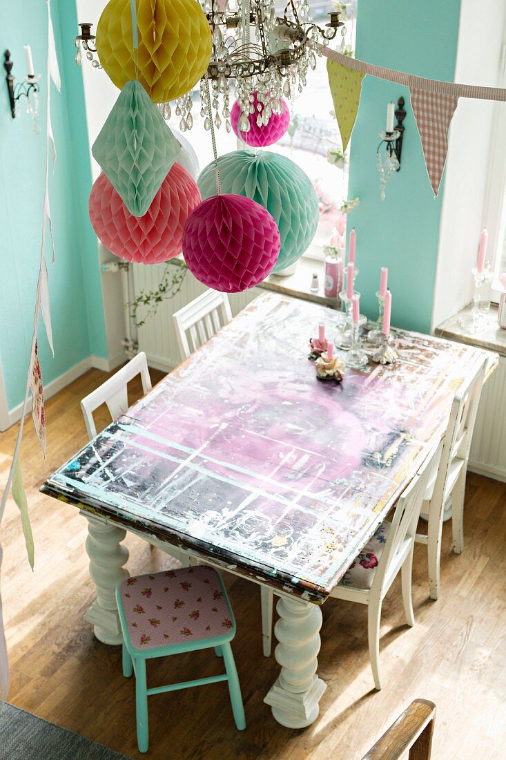 Table with various layers of paint and white wooden chairs below colourful paper pompoms