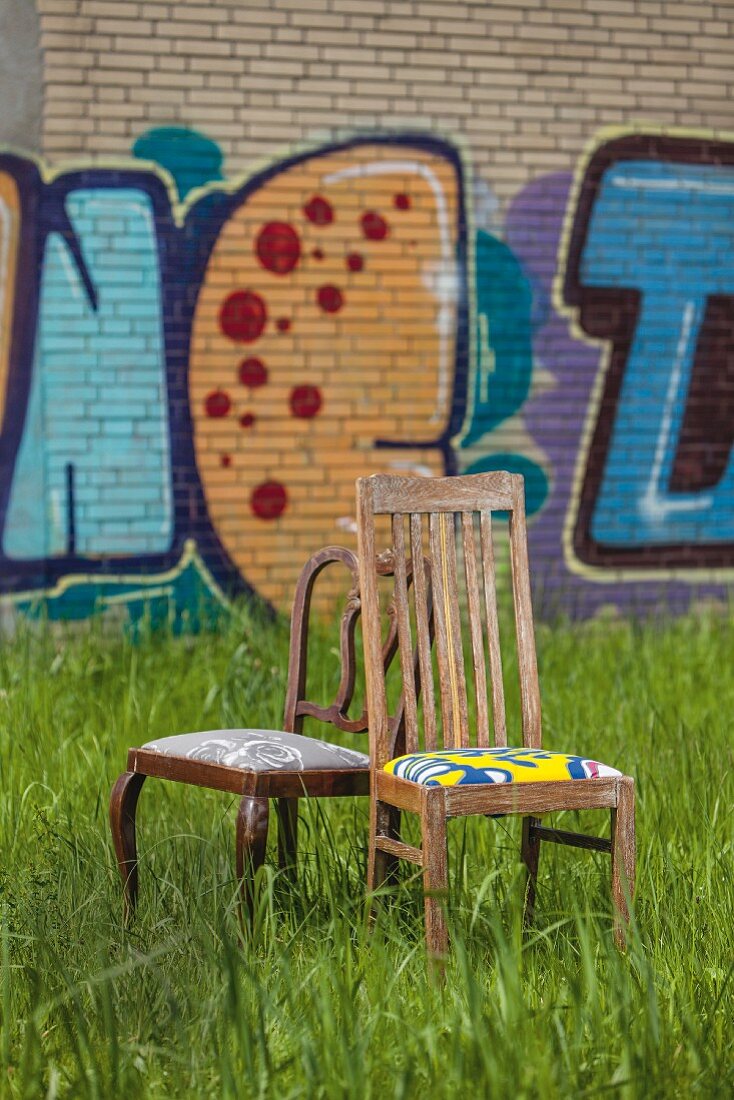 Alte Stühle mit neu bezogenen Sitzpolstern auf der Wiese, im Hintergrund Graffiti an Wand