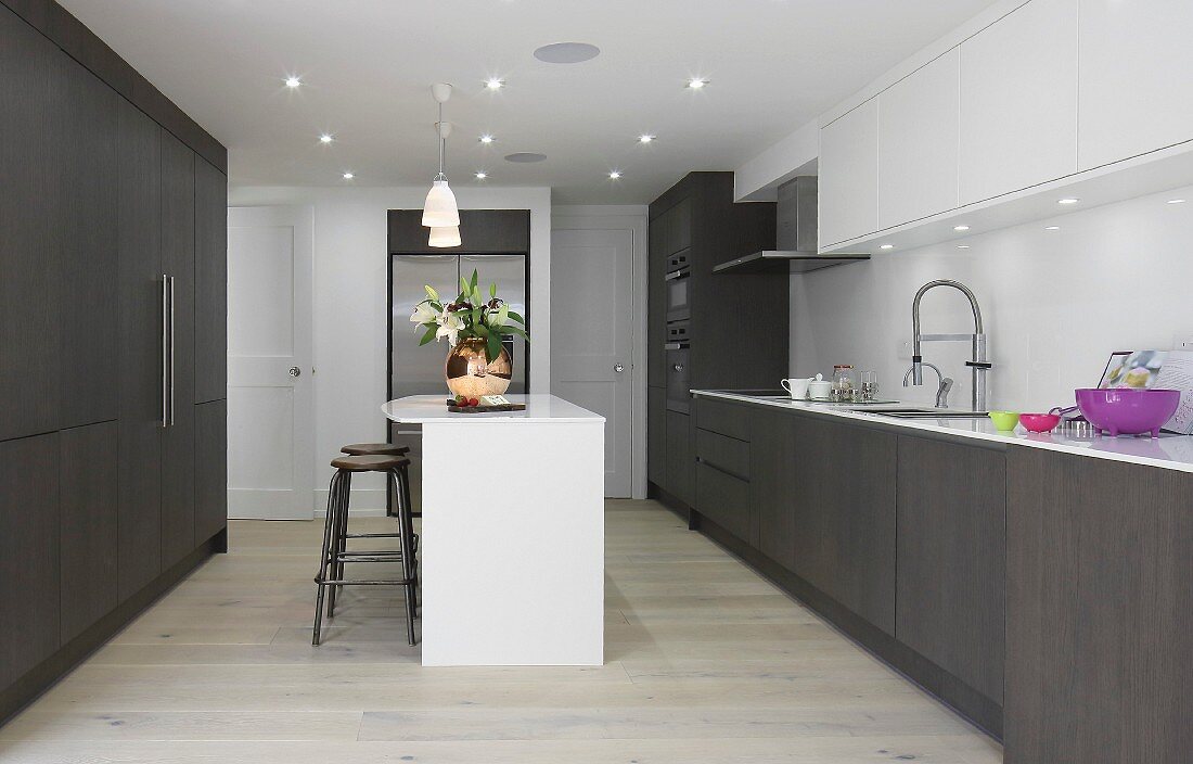 Einbauküche mit dunkelbraunen Holzfronten und weisser Kücheninsel, Deckeneinbaustrahler und weiße Hängeschränke in elegantem Ambiente