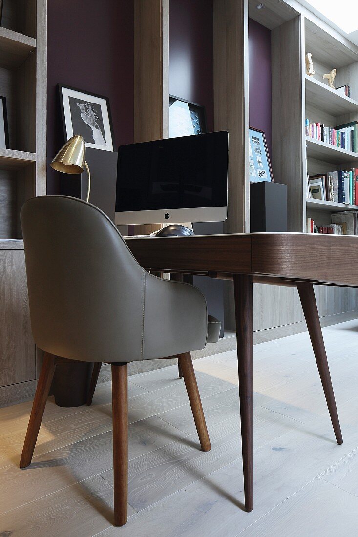 Eleganter Schreibtisch mit Monitor und gepolstertem grauem Lederstuhl im Retrostil neben raumhohem Regalelement