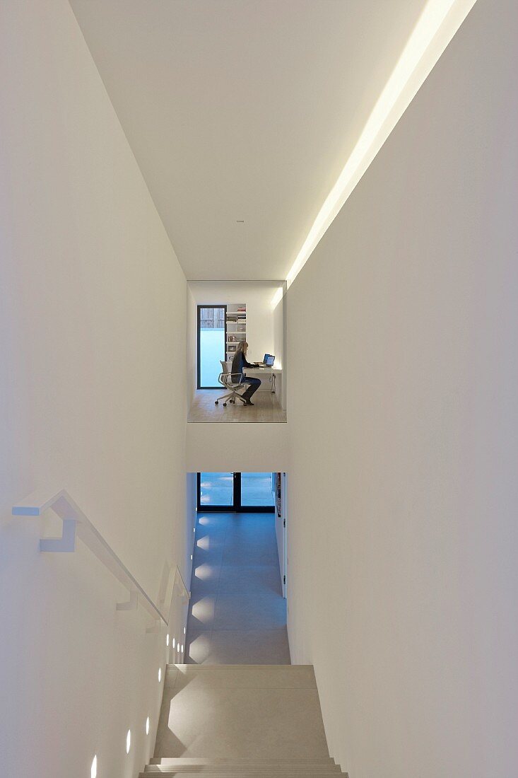 Blick von oben auf Treppenlauf in schmalem Treppenhaus mit indirekter Beleuchtung, im Hintergrund Blick durch Glaswand auf Frau am Schreibtisch
