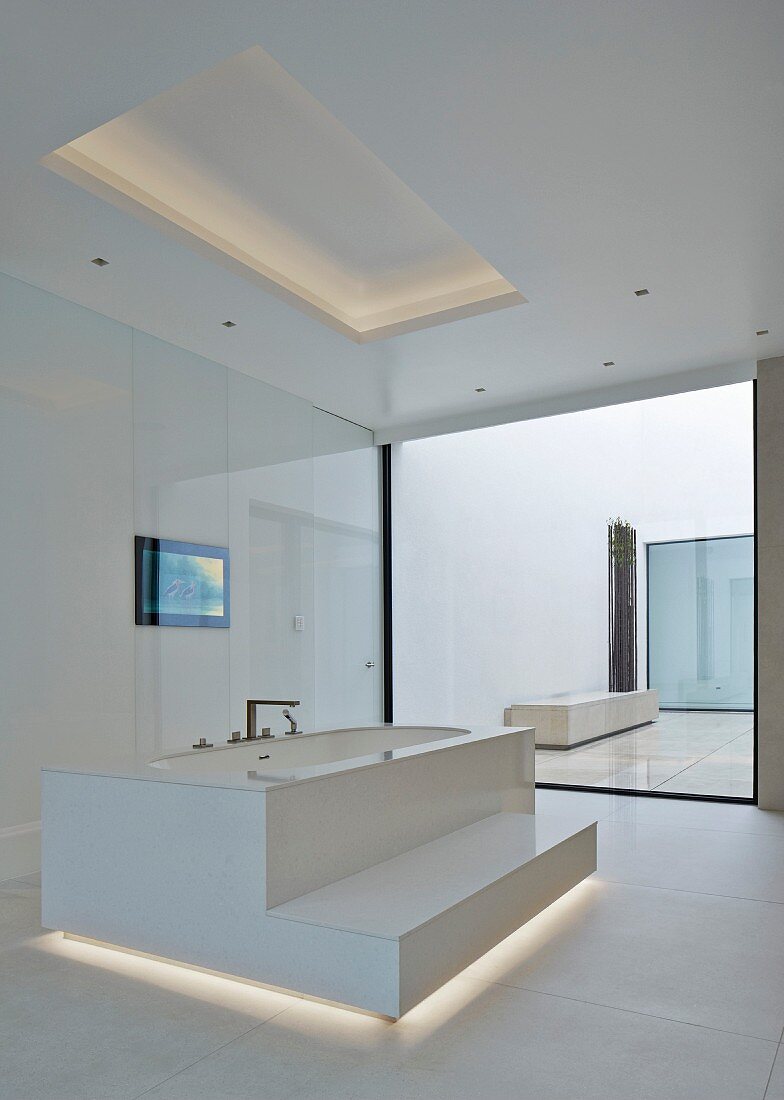 Freistehende Badewanne mit Stufe, in weißem, minimalistischem Designerbad mit indirekter Beleuchtung im Boden- und Deckenbereich,