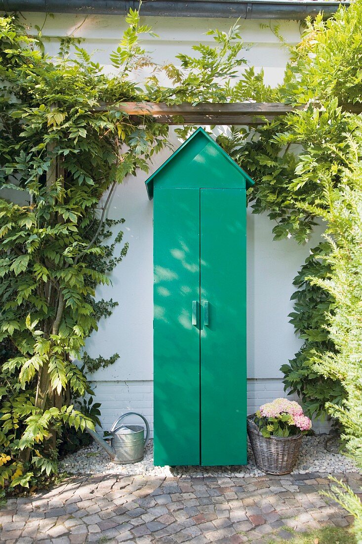 Closed green garden cupboard with gable roof below climber-covered pergola