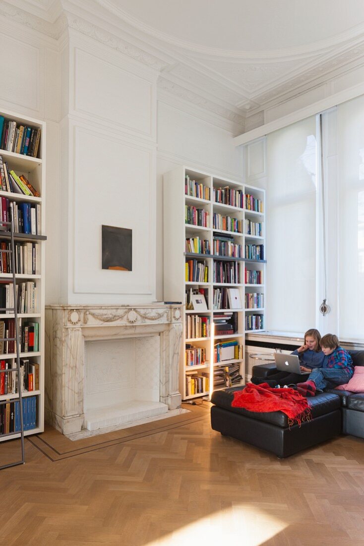 Wohnzimmerecke mit Stuckdecke, Kinder am Laptop auf schwarzem Sofa neben Bücherregal in Altbauwohnung