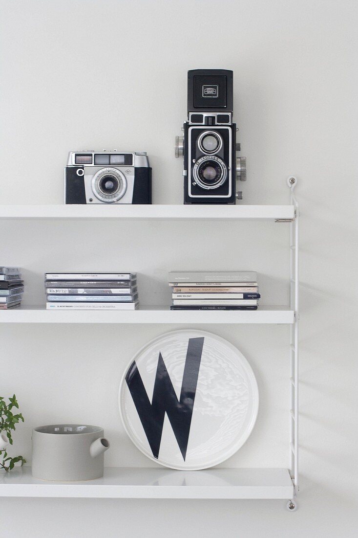 Retro-Fotoapparate und CD-Hüllen auf weißem String-Regal mit schwarz-weißem Wandteller