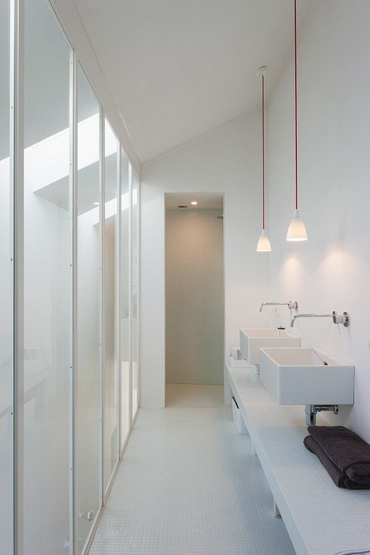 Schmaler weißer Waschbereich mit zwei getrennten Waschbecken und Pendelleuchten, gegenüber raumhohe Glaspaneele
