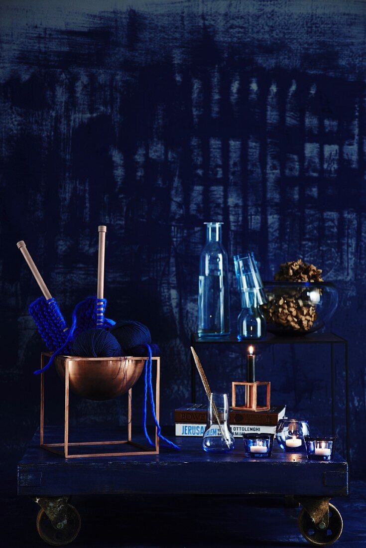 Trendfarben Blau und Kupfer - Kupferschale mit blauem Strickzeug und Glasware auf Holzbrett mit Rollen