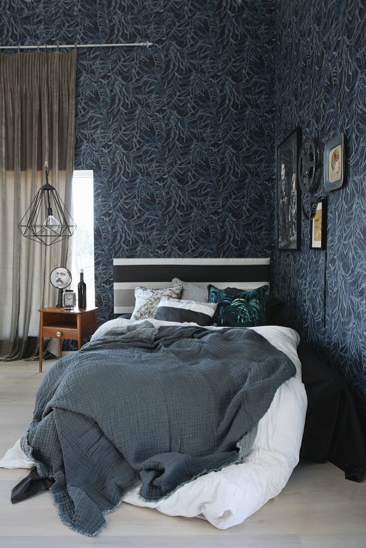 Bett mit grauer Tagesdecke und weisser Bettwäsche in maskuliner Zimmerecke mit floraler Mustertapete