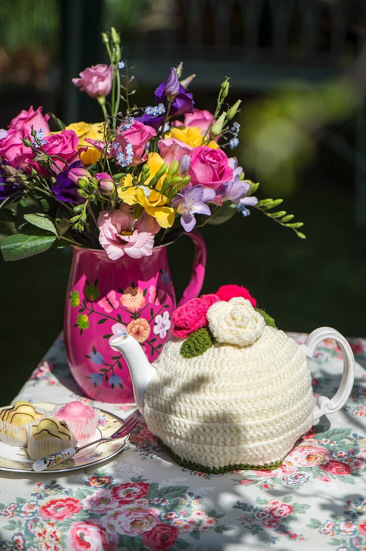 Bunter Blumenstrauss, Petit Fours und Teekanne mit gehäkeltem Kannenwärmer auf geblümter Tischdecke