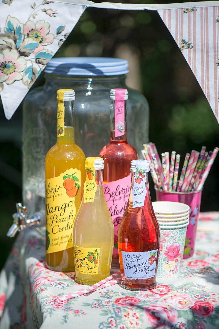 Verschiedene Saftflaschen und Pappbecher mit Trinkhalmen auf sonnigem Gartentisch mit geblümter Tischdecke