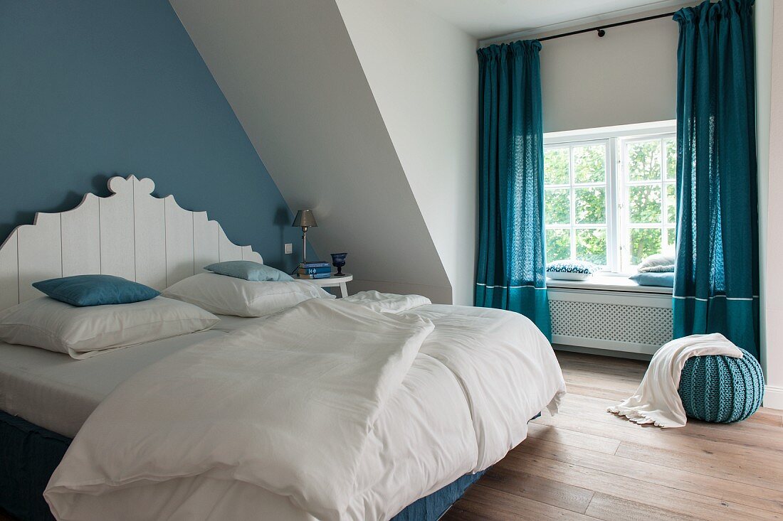 Schlafzimmer mit blauen Farbakzenten, Doppelbett mit weißem, geschnitztem Kopfteil vor blau getönter Wand, an Fenster blaue, bodenlange Vorhänge