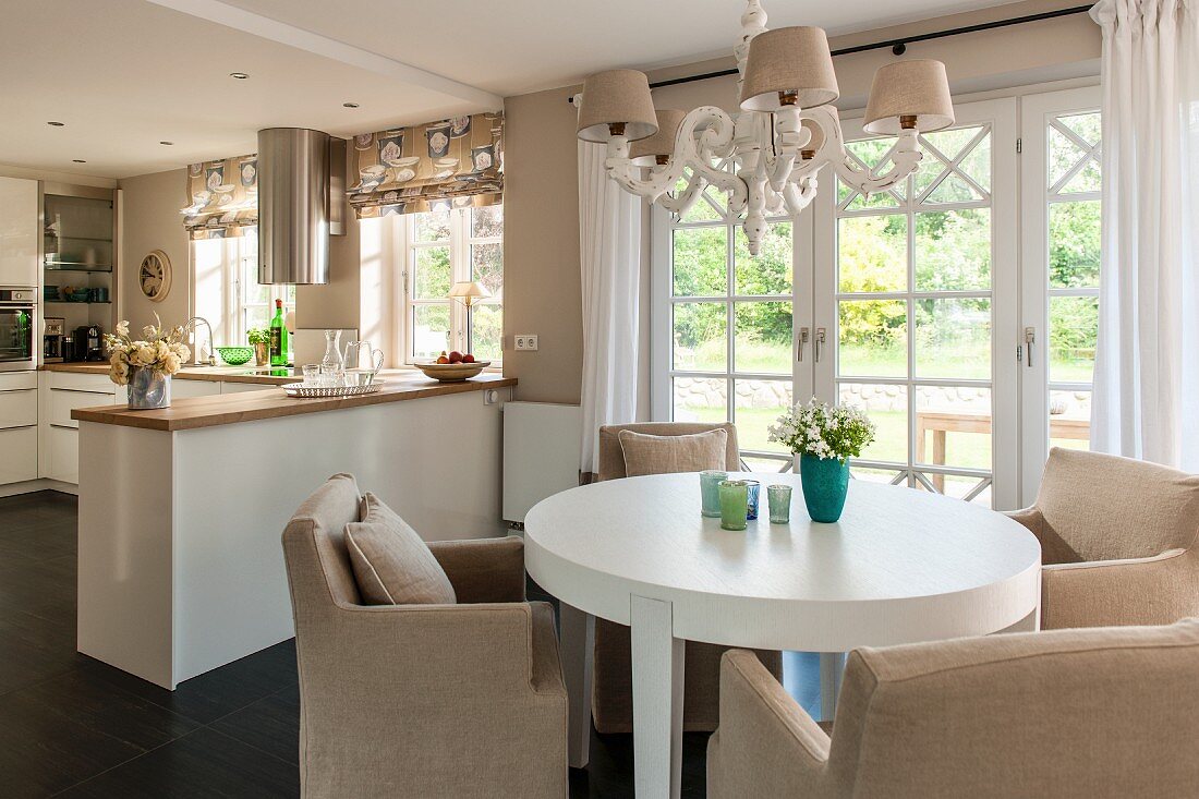 Runder Esstisch in Weiß und helle Polstersessel unter Kronleuchter, im Hintergrund offene Küche