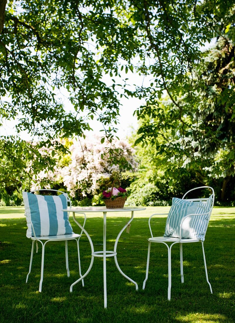 Zwei weiße, filigrane Metallstühle mit Kissen um runden Tisch auf Rasen im Garten