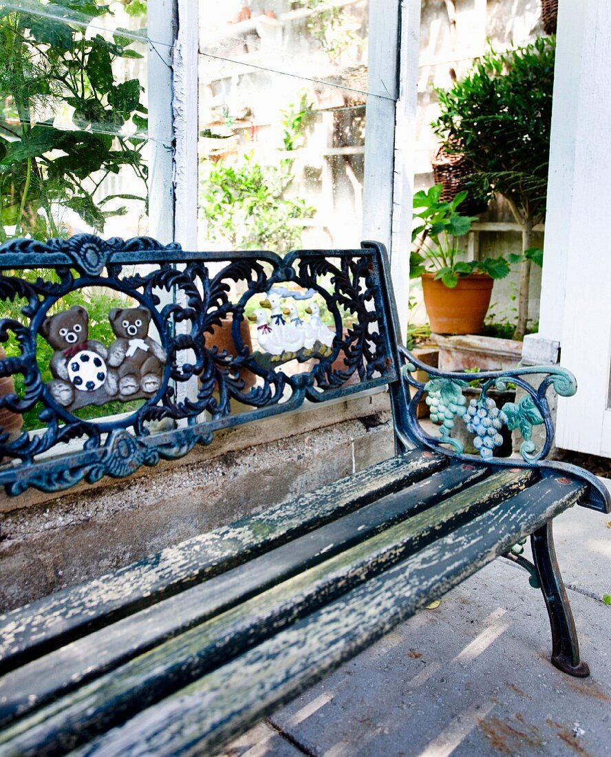 Vintage Gartenbank mit abblätternder Farbe und Rückenlehne aus verziertem Gusseisen vor Gewächshaus
