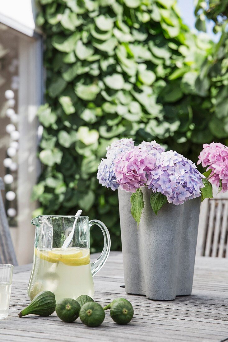 Lilafarbene und rosa Hortensien in grauer Vase, Erfrischungsgetränk in Glaskrug und frische Feigen auf Holztisch