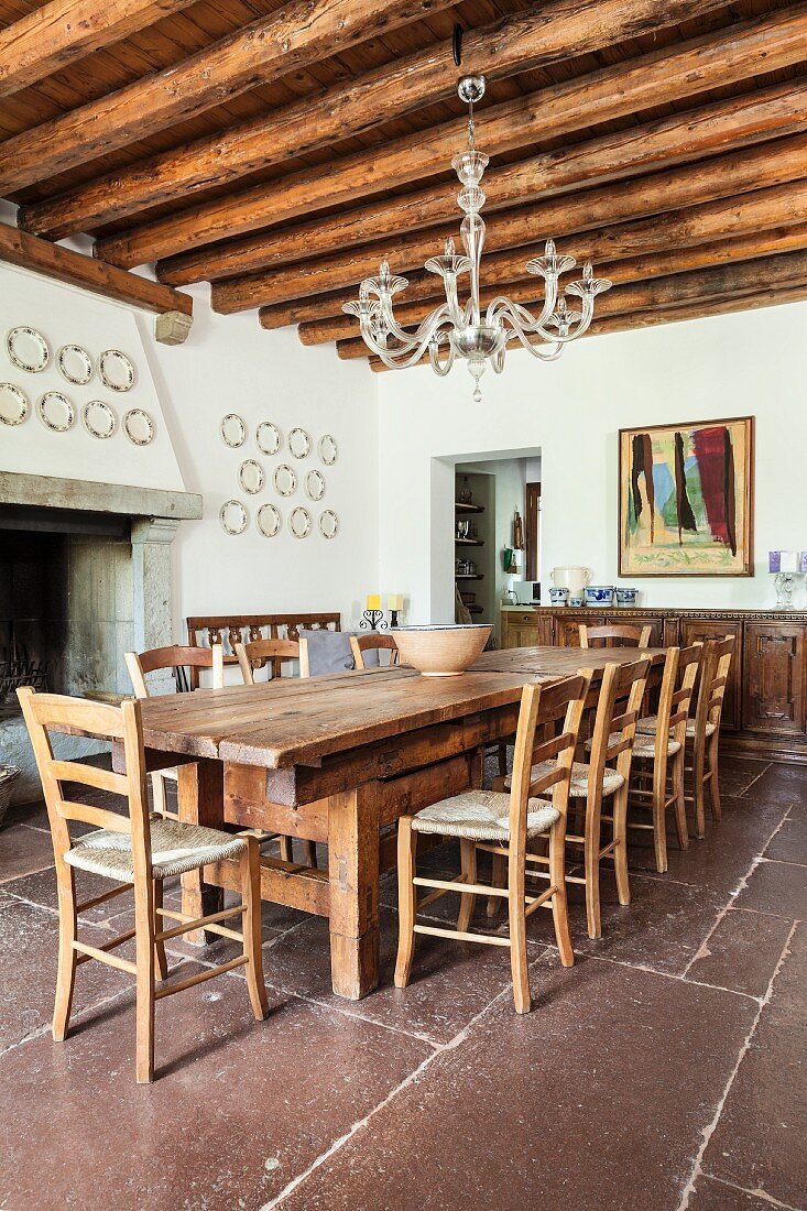Long wooden table and chairs on rustic stone floor below glass chandelier hung from wood-beamed ceiling in dining room