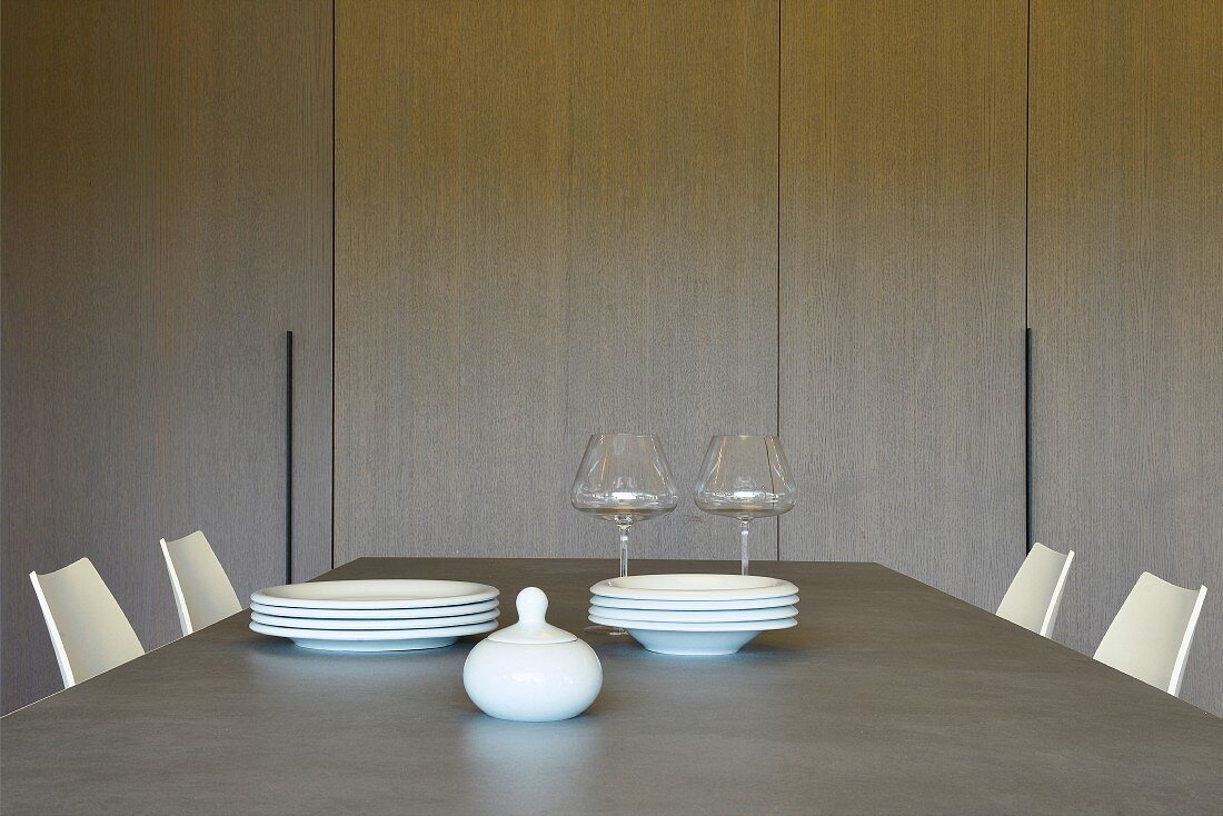 White sugar bowl, stacked plates and two wine glasses on dining table in front of fitted cupboards