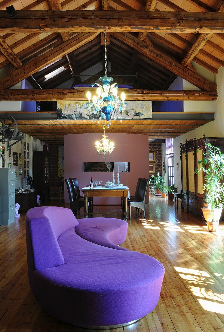 Geschwungenes violettes Sofa in Loftwohnung mit Holz-Dachstuhl und beleuchteten Kronleuchtern, im Hintergrund Essplatz unter Galerie
