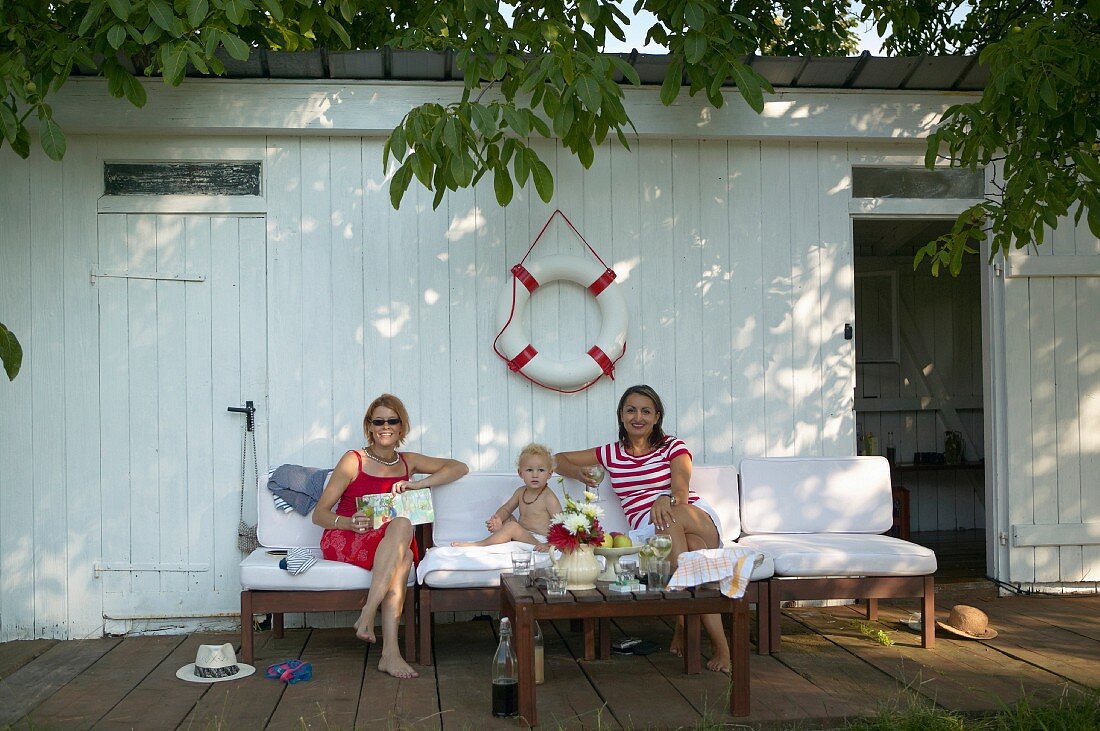 Two women and toddler on outdoor easy chairs below life belt on wall of white changing hut