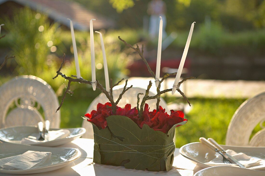 DIY-Kerzenhalter mit weissen Kerzen und Zweigen, roten Blüten und Blättern auf festlich gedecktem Tisch im Freien
