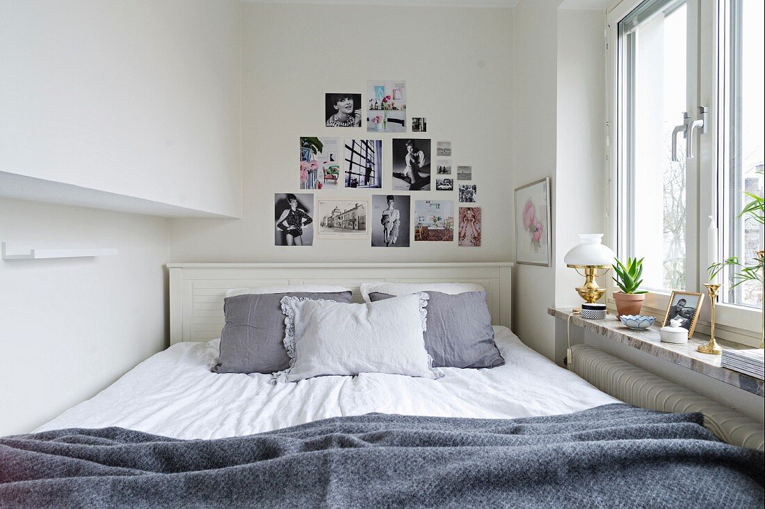 Doppelbett mit grauen Kissen an weiss lackiertem Kopfteil, darüber feminine Modefotografien dekorativ an weißer Wand neben Fenster