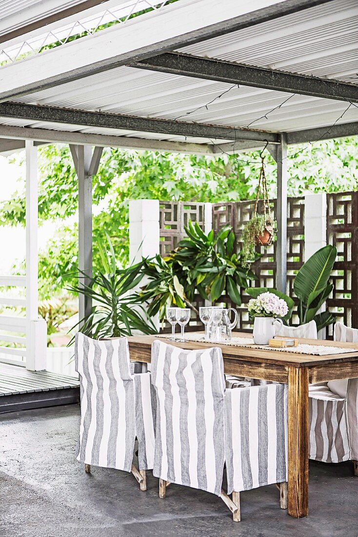 Covered terrace with gray and white striped armchairs and green plants in front of a privacy screen