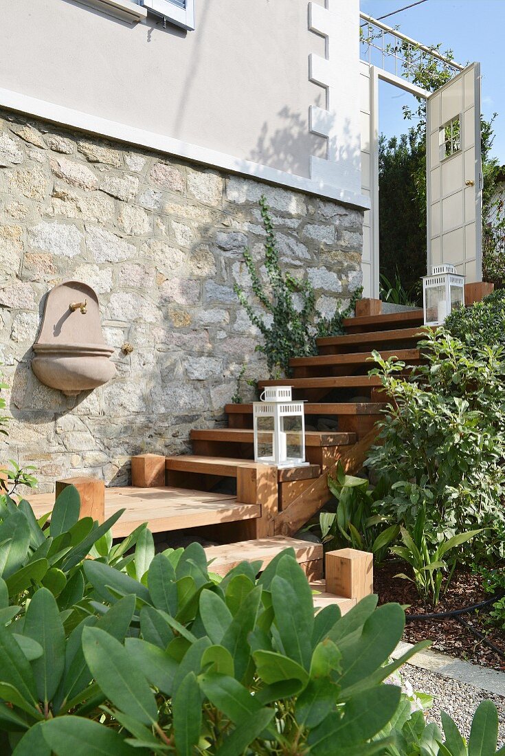 Gartenwaschbecken an Natursteinwand eines renovierten Landhauses mit Zedernholztreppe und aufgestellten weissen Laternen