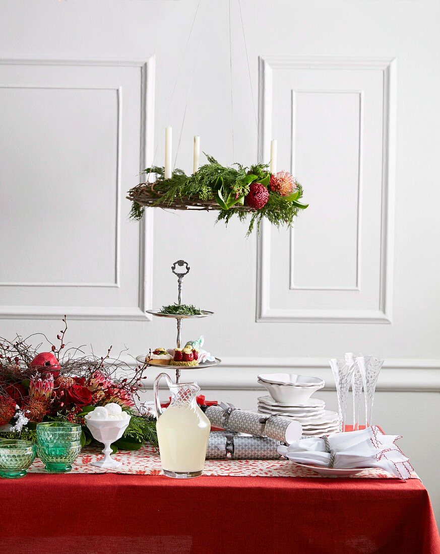Weihnachtlich geschmückter Tisch mit roter Tischdecke und weißem Geschirr, oberhalb aufgehängter Adventskranz