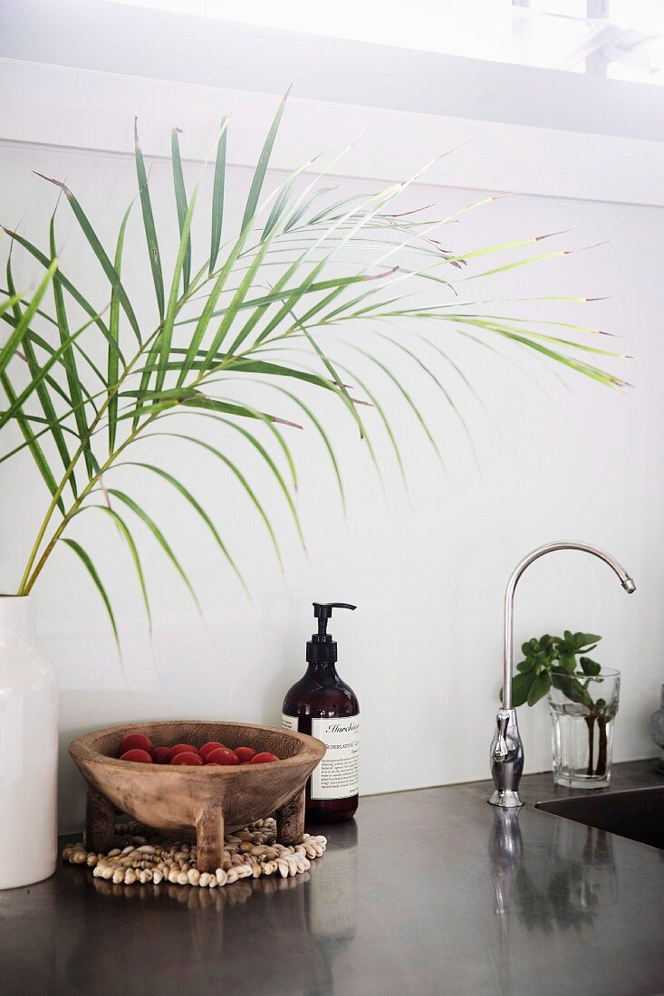 Küchenarbeitsplatte mit grau polierter Oberfläche, Palmenzweige in Vase und rustikale Holzschale neben Seifenspender am Spülbecken