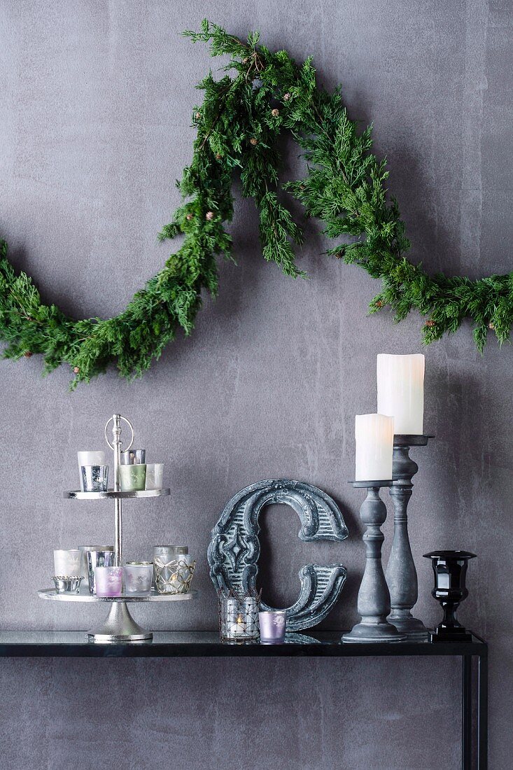 Weihnachteliche Zweig-Girlande an Wand über Dekoarrangement mit Teelichtgläsern und Kerzenständern
